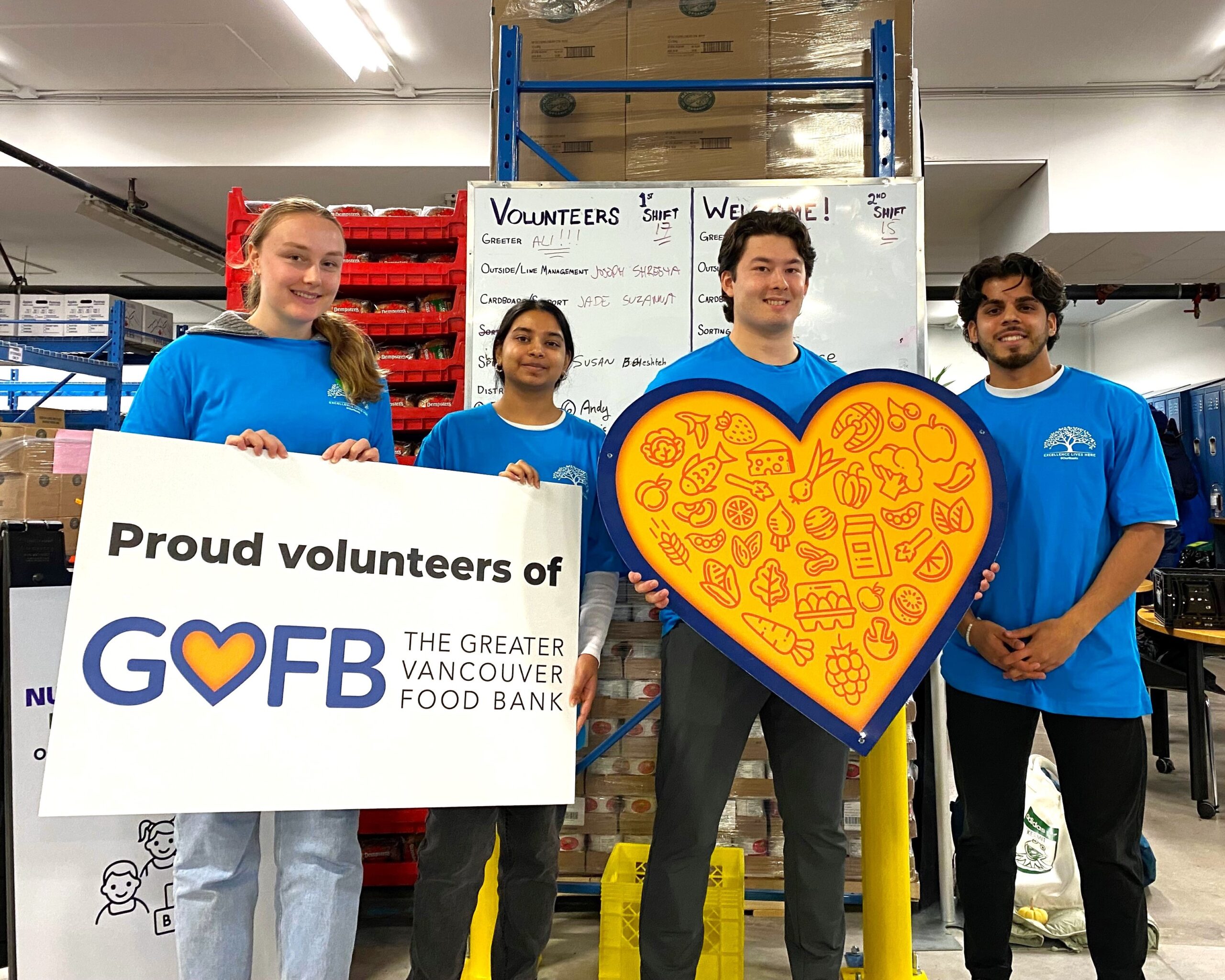 Four people standing. One holds a Greater Vancovuer Food Bank sign and another holds a large, yellow heart.